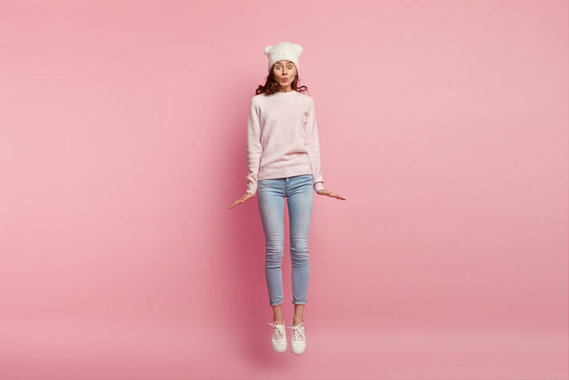 灵感可爱女孩的照片在粉色空间的空中跳跃 戴着帽子 毛衣 牛仔裤和运动鞋 嘴唇一直叠着凝视自由情绪