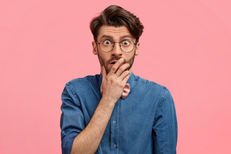 鬃毛惊呆了的帅哥捂着嘴 瞪着眼睛 透过眼镜 穿着牛仔衬衫惊奇手掌表情