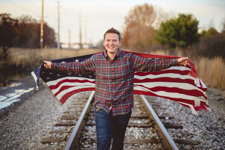 日微笑的男性手持美国国旗在火车轨道上行走美国爱国主义步行