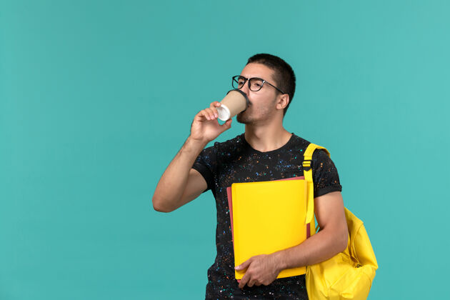 饮酒男学生穿着深色t恤 背着黄色背包 拿着不同的文件 在蓝色的墙上喝着咖啡大学歌手音乐家