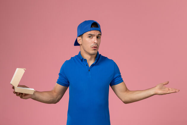 粉色正面图身穿蓝色制服披肩的年轻男性快递员拿着粉色墙上的小空快递食品包男人制服食物
