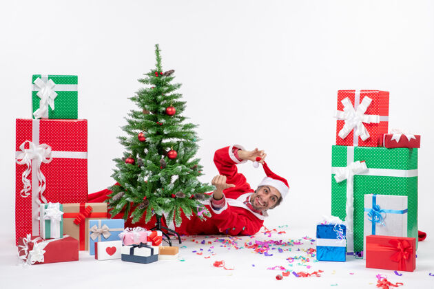 说谎节日气氛与年轻积极的圣诞老人躺在圣诞树后靠近礼物白色背景节日年轻圣诞老人