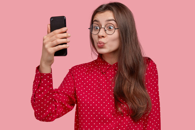 眼镜美丽迷人的女人在手机上自拍 在镜头下撅着嘴 戴着圆眼镜 享受闲暇时光波尔卡多制造鬼脸