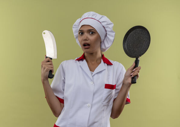 切肉刀惊讶的年轻厨师女身穿厨师制服拿着煎锅和切肉刀煎炸平底锅惊讶