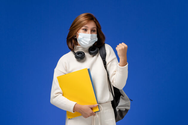 女人正面图身穿白色球衣的年轻女学生戴着面具 背着背包 拿着文件在蓝色的墙上欢呼雀跃大学背包风景