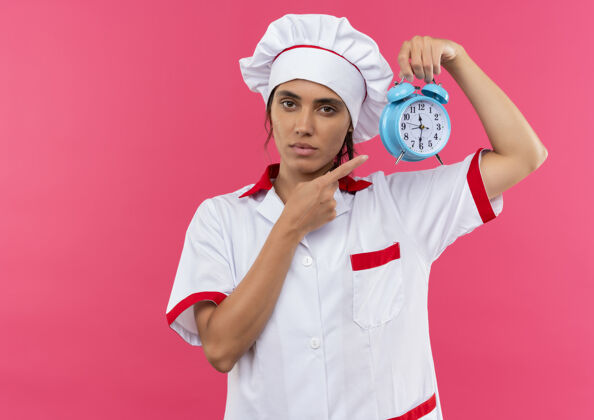 女年轻的女厨师穿着厨师制服 拿着手指头对着墙上的闹钟 墙上有一个单独的粉色空间钟穿拿