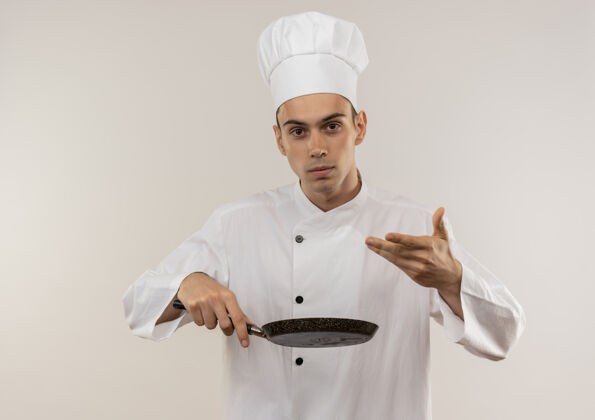 拿着年轻的男厨师穿着厨师制服拿着煎锅站在隔离的白色墙上厨师穿着看着