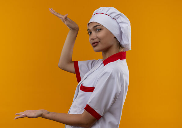 厨师令人高兴的年轻厨师女穿厨师制服显示在孤立的橙色墙壁与复制空间的大小制服年轻展示