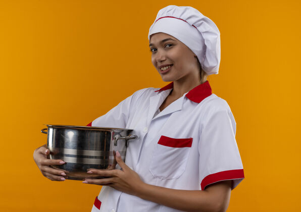 女微笑的年轻女厨师穿着厨师制服 拿着平底锅 背景是孤立的橙色制服厨师穿着