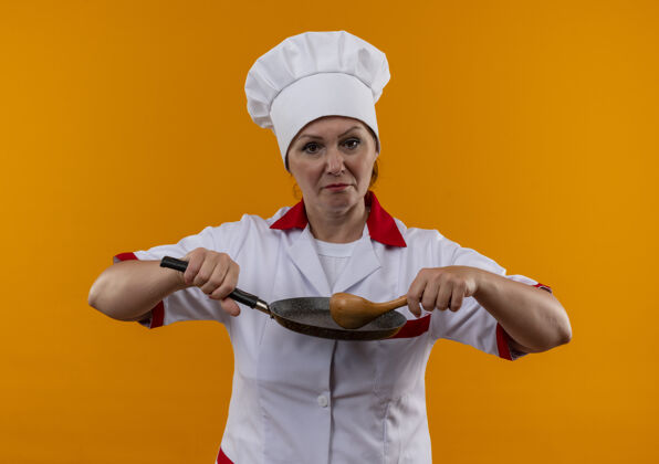 女自信的中年女厨师穿着厨师制服 拿着煎锅和勺子站在隔离的黄色墙上勺子制服厨师