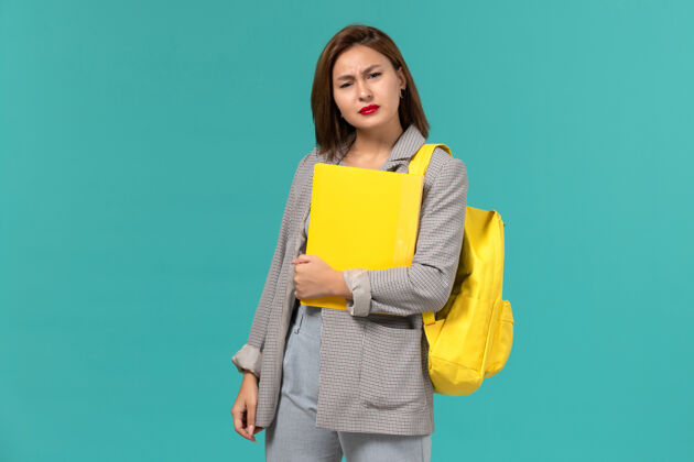 学校身穿灰色夹克 背着黄色背包 在浅蓝色墙上拿着文件的女学生的正视图灯光正面书籍