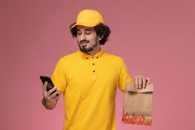 男人正面图身穿黄色制服的男快递员拿着食品包在粉色墙上打电话视图谈话电话