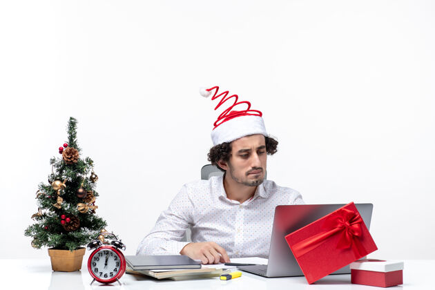 商人快乐积极的年轻商人戴着有趣的圣诞老人帽子在白色背景的办公室里仔细检查自己圣诞节圣诞老人电脑