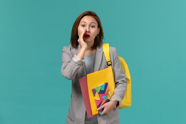 耳语身着灰色夹克 背着黄色背包 拿着文件和文案的女学生在蓝色墙上低语的正面照片学校夹克女孩