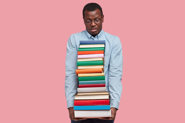不满不高兴的非裔美国科学家或学生背着厚厚的一堆教科书 穿着优雅的衬衫墙壁男性眼镜
