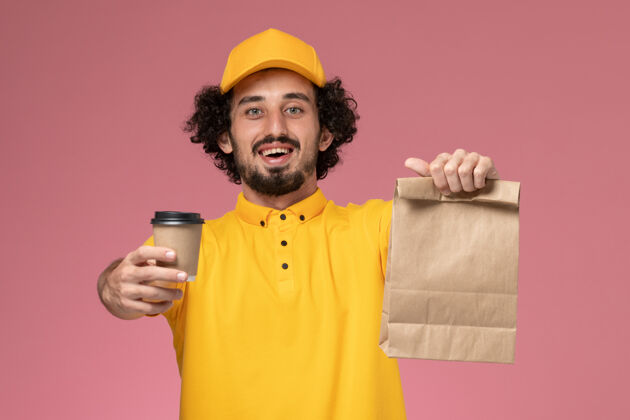 包裹正面图：身穿黄色制服 披风的男性快递员手持咖啡杯和食品包 放在粉红色的墙上帽子男食物