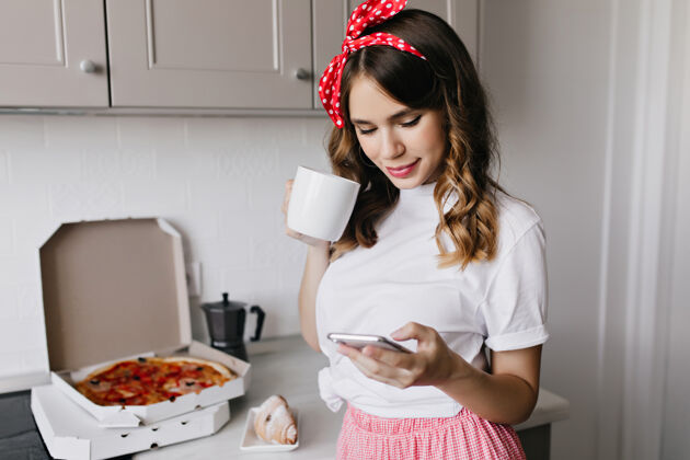 食物快乐的年轻女子在厨房里喝着咖啡 发着短信早餐时 欣喜若狂的卷发女模特在家摆姿势模特寒冷披萨