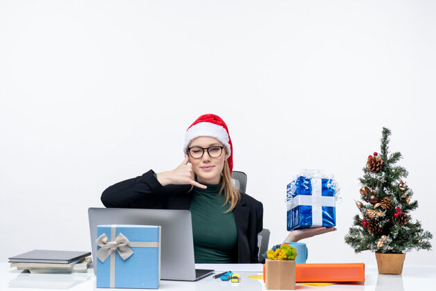 眼镜戴着圣诞老人帽戴着眼镜的年轻女子坐在桌子旁 在白色背景上展示圣诞礼物制作的“召唤我”手势 这是节日的气氛电话圣诞老人圣诞节