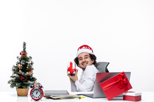 圣诞节带着微笑的积极的年轻商人 戴着圣诞老人的帽子 坐在办公室里 在白色的背景上愉快地指着他的礼物 新年的气氛快乐商人办公室