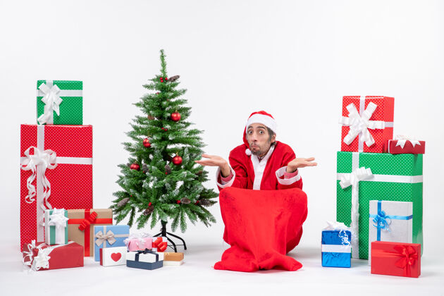 圣诞树困惑的年轻人打扮成圣诞老人 拿着礼物和装饰过的圣诞树坐在地上的白色背景上人年轻人地