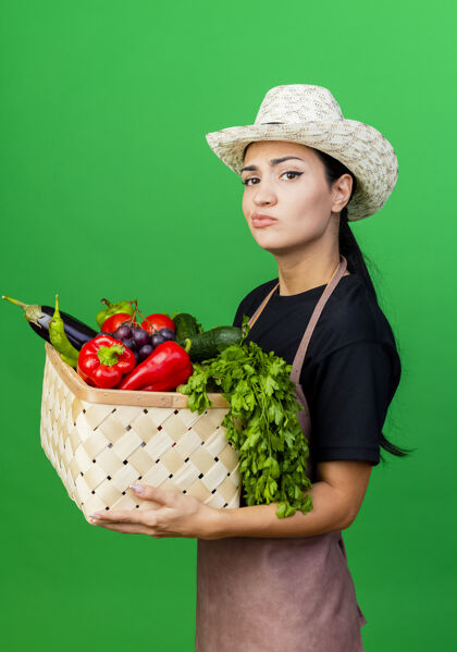 绿色年轻漂亮的女园丁 围着围裙 戴着帽子 手里拿着装满蔬菜的篮子 带着怀疑的表情园丁帽子女人