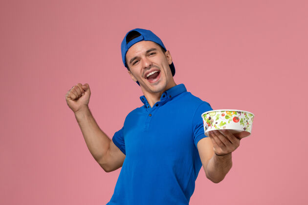 人正面图身穿蓝色制服斗篷的年轻男性信使手持圆形的投递碗 在浅粉色的墙上欢呼雀跃粉色喜悦碗