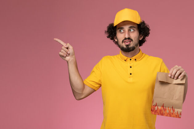正面正面图穿着黄色制服的男快递员拿着浅粉色墙上的纸质食品包工人送货帽子