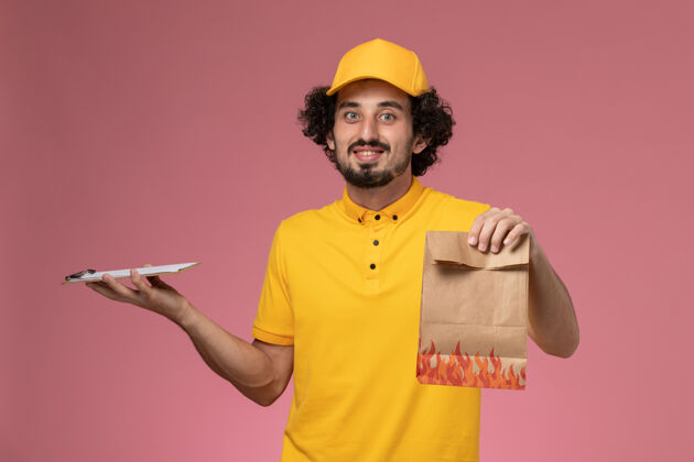 食物正面图身穿黄色制服的男快递员拿着食品包和笔记本放在粉红色的墙上制服粉色快递