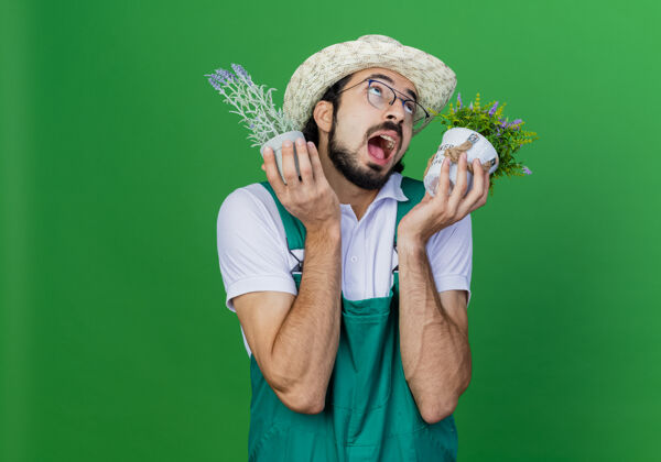 胡须年轻的留着胡须的园丁 穿着连体衣 戴着帽子 手里拿着盆栽植物 高兴而兴奋地抬起头来兴奋穿着男人