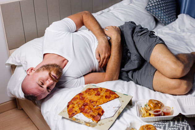 卧室一个留着大胡子的白种人在吃过多的快餐比萨饼和汉堡后躺在床上吃披萨家