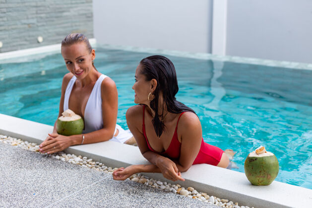 人两位亚洲人和白种人的女性朋友在别墅的游泳池化妆椰子一起放松