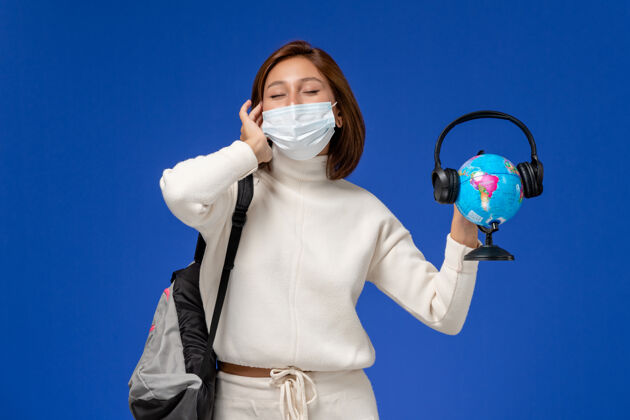 女人正面图身穿白色球衣的年轻女学生戴着面具 背着背包 拿着地球仪 蓝色墙上挂着耳机年轻的女学生大学地球