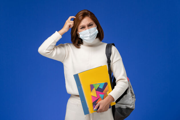 女性正面图穿着白色运动衫的年轻女学生戴着带包的面具 在蓝色的墙上思考着抄写本专业成人女人