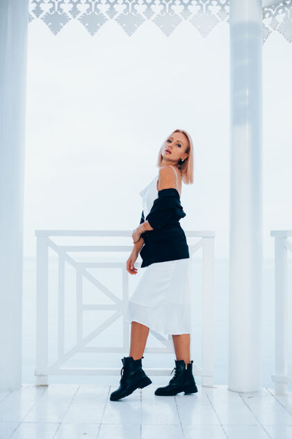 时尚时尚写真时尚的白种女人在丝绸长裙黑色外套和大靴子在豪华的地方与海景站立金发女性