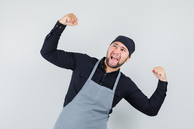 餐厅男厨师穿着衬衫 围裙 展现获胜者的姿态 看起来很幸福 正面视图烹饪展示准备