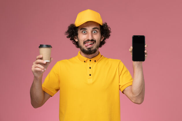 工人正面图：身穿黄色制服 披风的男性快递员 在粉色墙上拿着送货咖啡杯和电话制服人查看