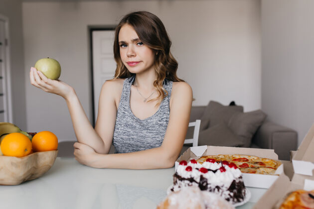 选择沉思的高加索女孩想着她的饮食 手里拿着苹果一张严肃的卷发女人拿着健康的食物和比萨饼摆在桌子前的室内照片早餐快乐金发