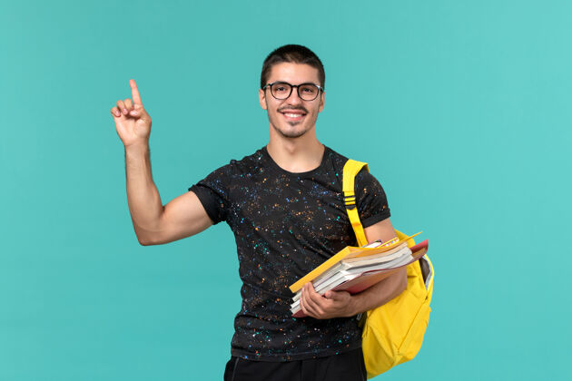 学生身穿深色t恤黄色背包的男学生正面图 浅蓝色墙上有文件和书籍大学教训大学