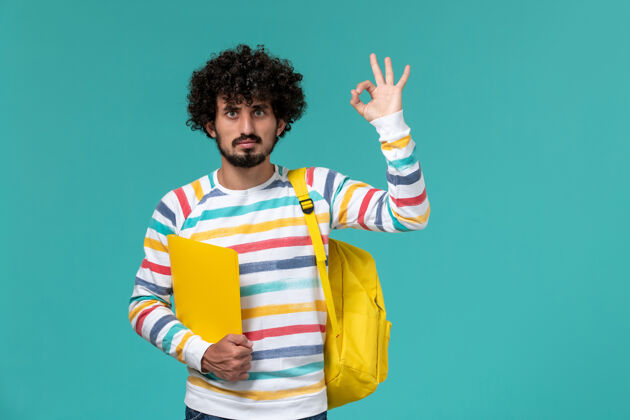 衬衫身穿条纹衬衫 背着黄色背包 在蓝色墙上拿着文件的男学生的正视图人孩子微笑