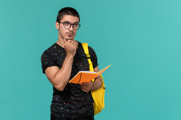 大学身穿深色t恤黄色背包的男学生正面图拿着抄写本在蓝色墙上思考持有漂亮大学