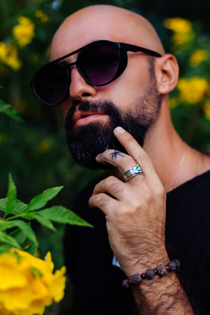 时尚在公园里 一个戴着太阳镜 皮肤黝黑 留着胡须的野蛮男子 手指架上有棕榈树纹身 周围是黄色的花朵花园情感户外