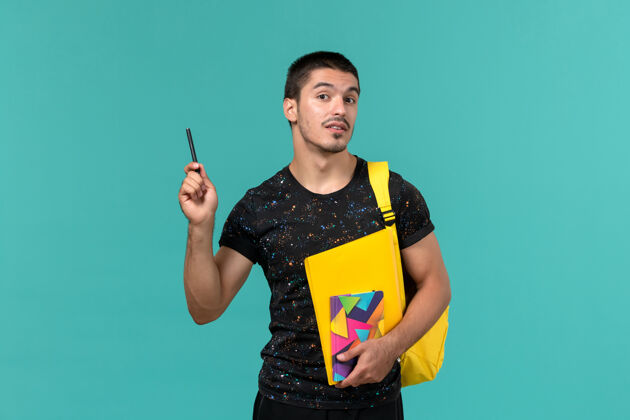 学校前视图中的男学生穿着深色t恤黄色背包拿着文案笔和文件在浅蓝色的墙上教训人成人