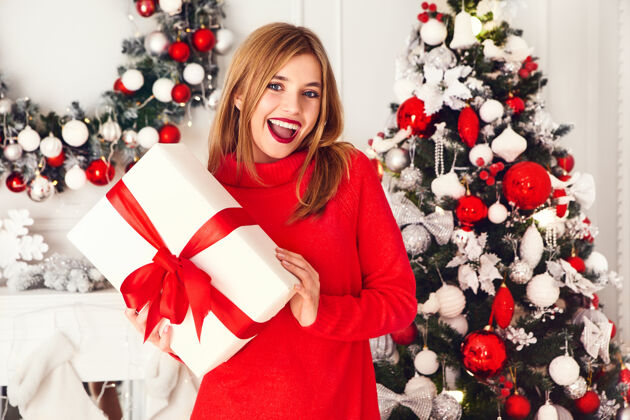 装饰微笑的女人拿着许多礼品盒在装饰好的圣诞树旁摆姿势毛衣惊喜青少年