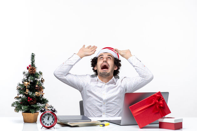办公室愤怒紧张的年轻商人 戴着滑稽的圣诞老人帽子 在白色背景的办公室里指着上方紧张圣诞老人年轻