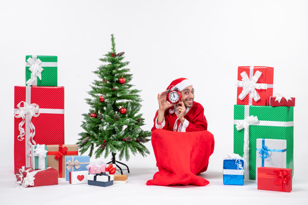 圣诞老人喜怒无常的圣诞老人坐在地上 展示着钟表附近的礼物和装饰圣诞树上的白色背景股票照片圣诞树股票礼物