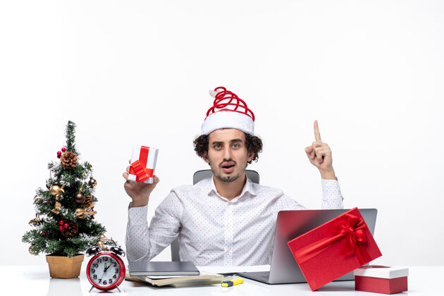 礼物圣诞气氛与微笑的年轻商人圣诞老人帽子和举行他的礼物指向上方的白色背景年轻商人年轻圣诞老人
