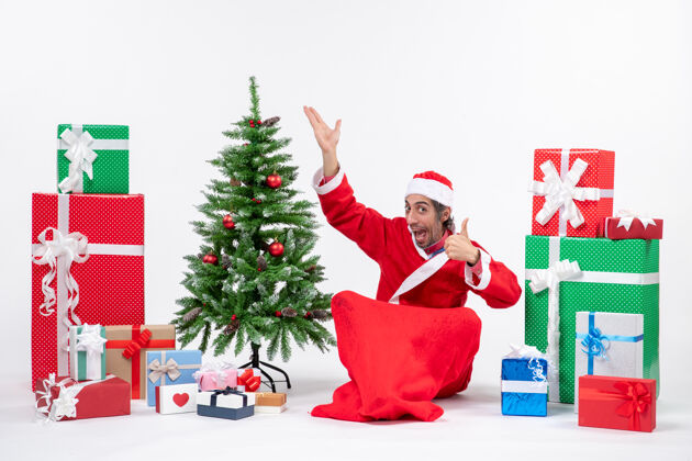 礼物有趣的年轻人打扮成圣诞老人与礼物和装饰圣诞树坐在地上指着上面作出完美的姿态在白色背景地面圣诞老人坐着