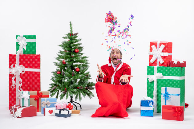 圣诞老人喜庆的节日气氛与有趣的积极惊喜圣诞老人坐在地上 玩圣诞装饰品附近的礼物和装饰圣诞树上的白色背景圣诞老人坐着节日