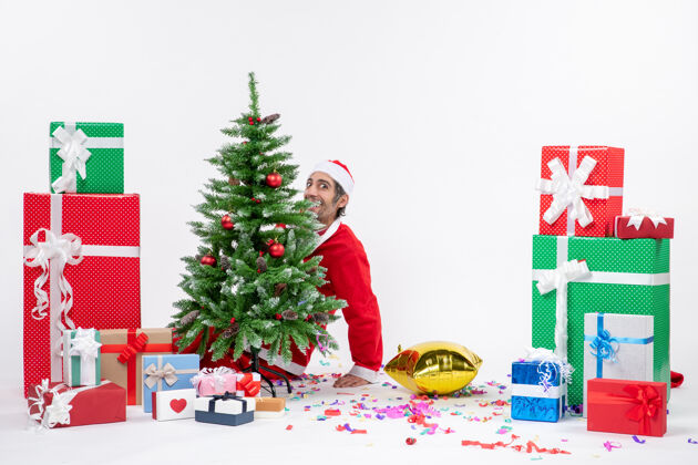 礼物圣诞气氛与年轻积极有趣的圣诞老人躺在圣诞树后 在白色背景上不同颜色的礼物附近不同沙拉圣诞