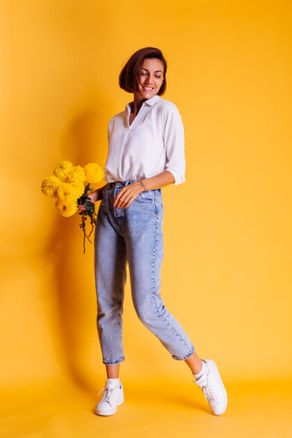 微笑摄影棚拍摄的黄色背景快乐的白人妇女短发穿着休闲服白衬衫和牛仔裤手持一束黄色紫苑积极礼物年轻